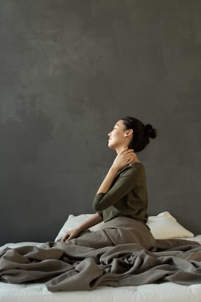 Side view of a woman sitting on a bed indoors, massaging her neck before starting stress free morning.